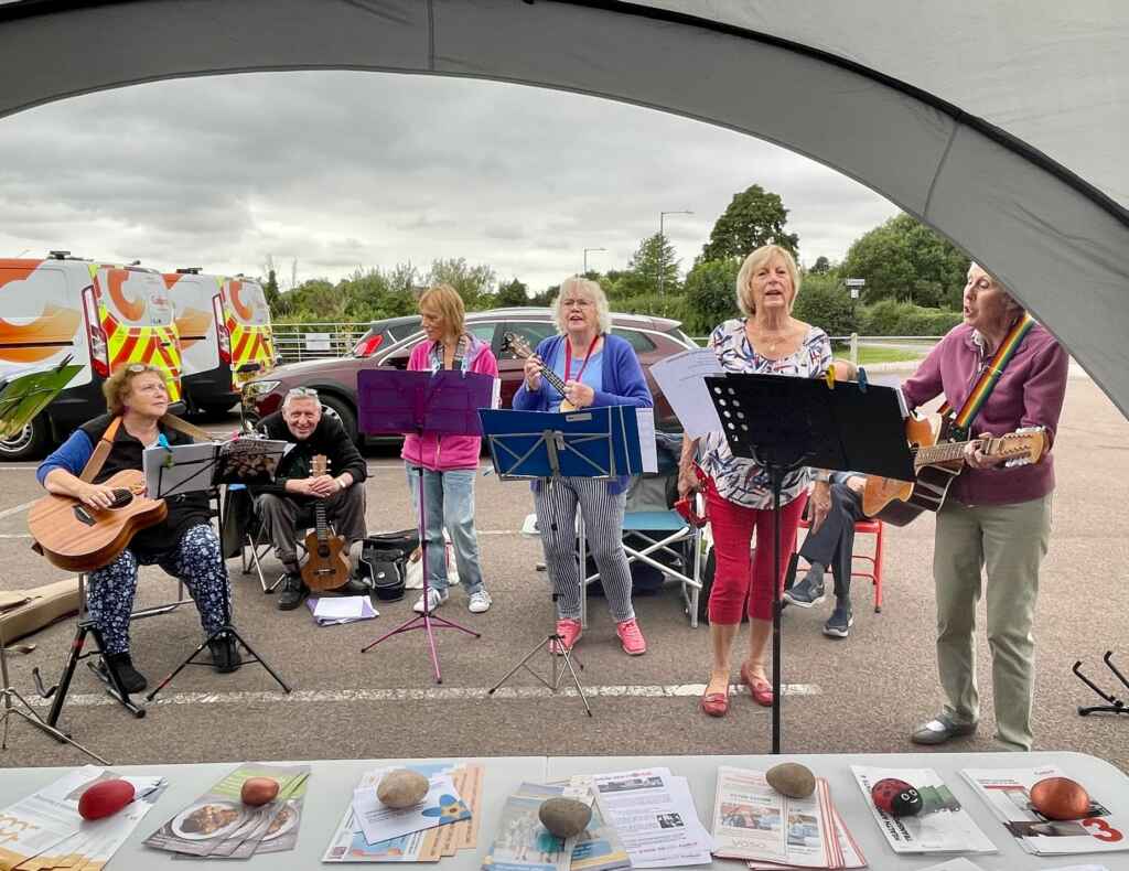 A band playing instruments and singing under a large canopy, with vans and cars in the background