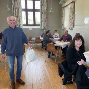 A man standing in a village hall holding a carrier bag with takeaway breakfasts with people sitting in the background