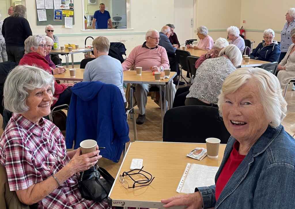 Two women drinking coffee at a table in front of other people sitting in a village hall