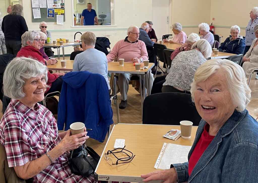 Two women drinking coffee at a table in front of other people sitting in a village hall