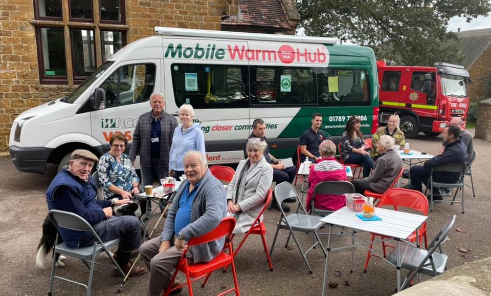 A group of people sitting at picnic tables and chairs with a green and white minibus in the background with the branding "Mobile Warm Hub" and part of a red fire engine visible in the far background