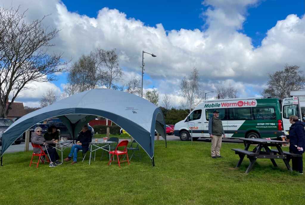 A large gazebo with people sitting underneath next to a picnic bench, with a green and white minibus with "Mobile Warm Hub" branding in the background, on a grassy area