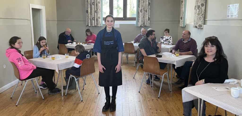 Young girl with a black apron standing in front of tables and chairs with people eating and drinking in a hall