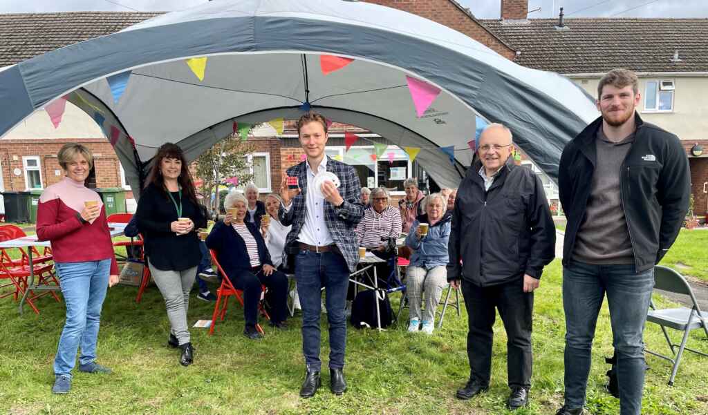 Young man holding cards with a small group of people standing and more people sitting behind him under a large gazebo with colourful bunting outside